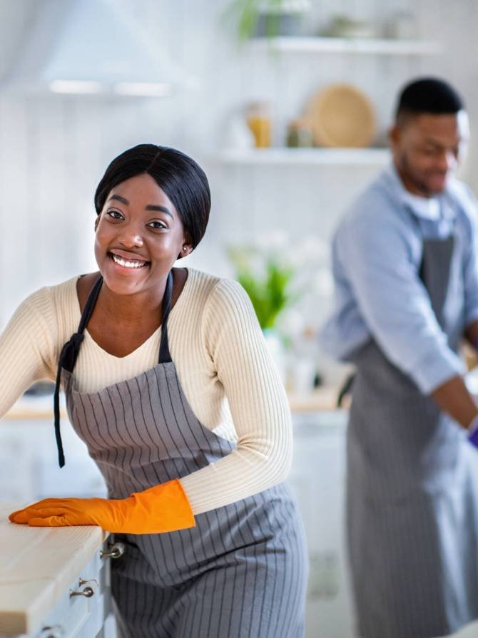 Portrait of smiling black lady in apron and rubber gloves wiping kitchen table, her boyfriend washing floor on background, blank space. Positive African American couple cleaning their house